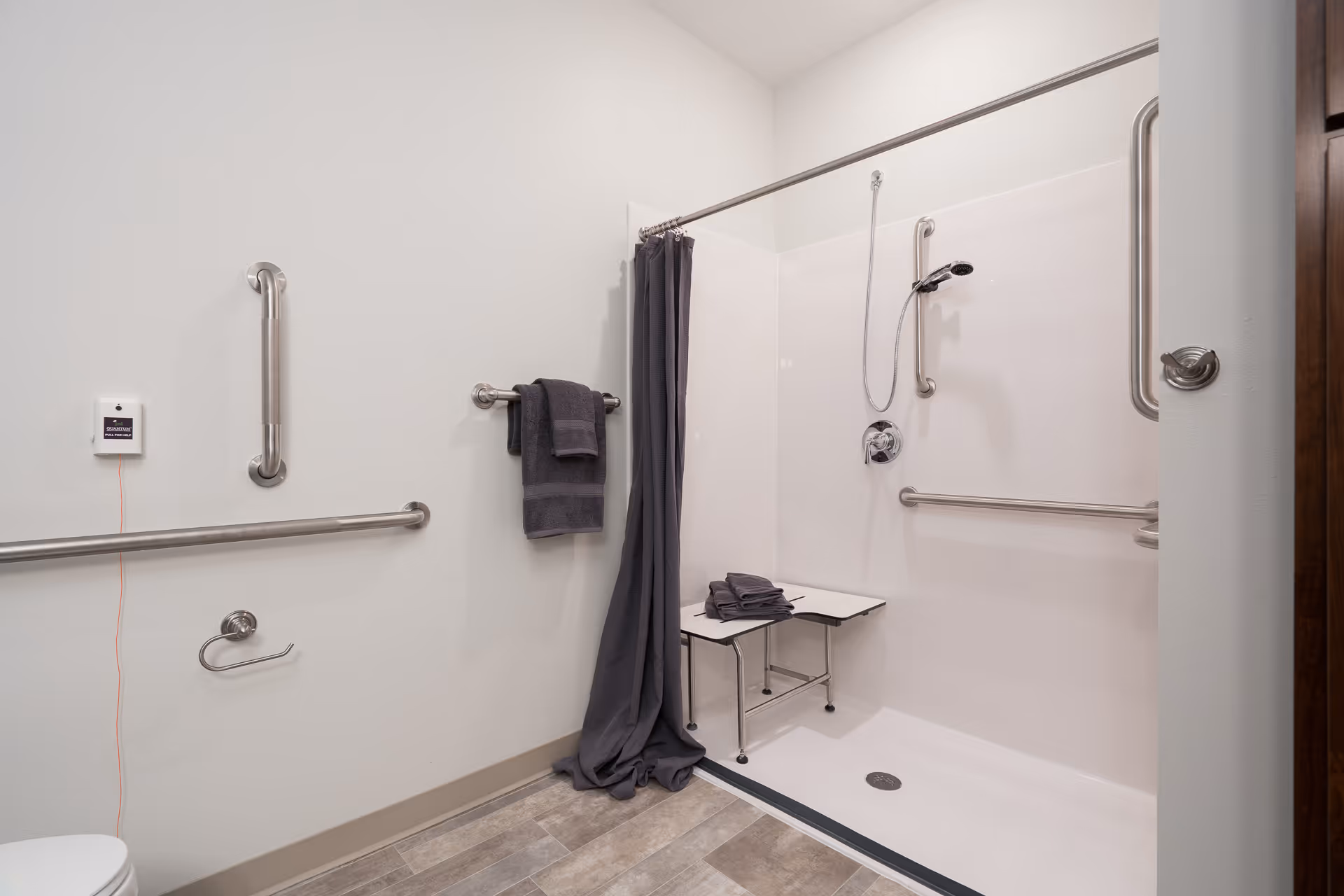 Accessible bathroom with a walk-in shower featuring grab bars, a fold-down shower seat, and a handheld showerhead. There are dark gray towels hanging on a towel rack and folded on the shower seat. The bathroom has light-colored walls and a tiled floor.
