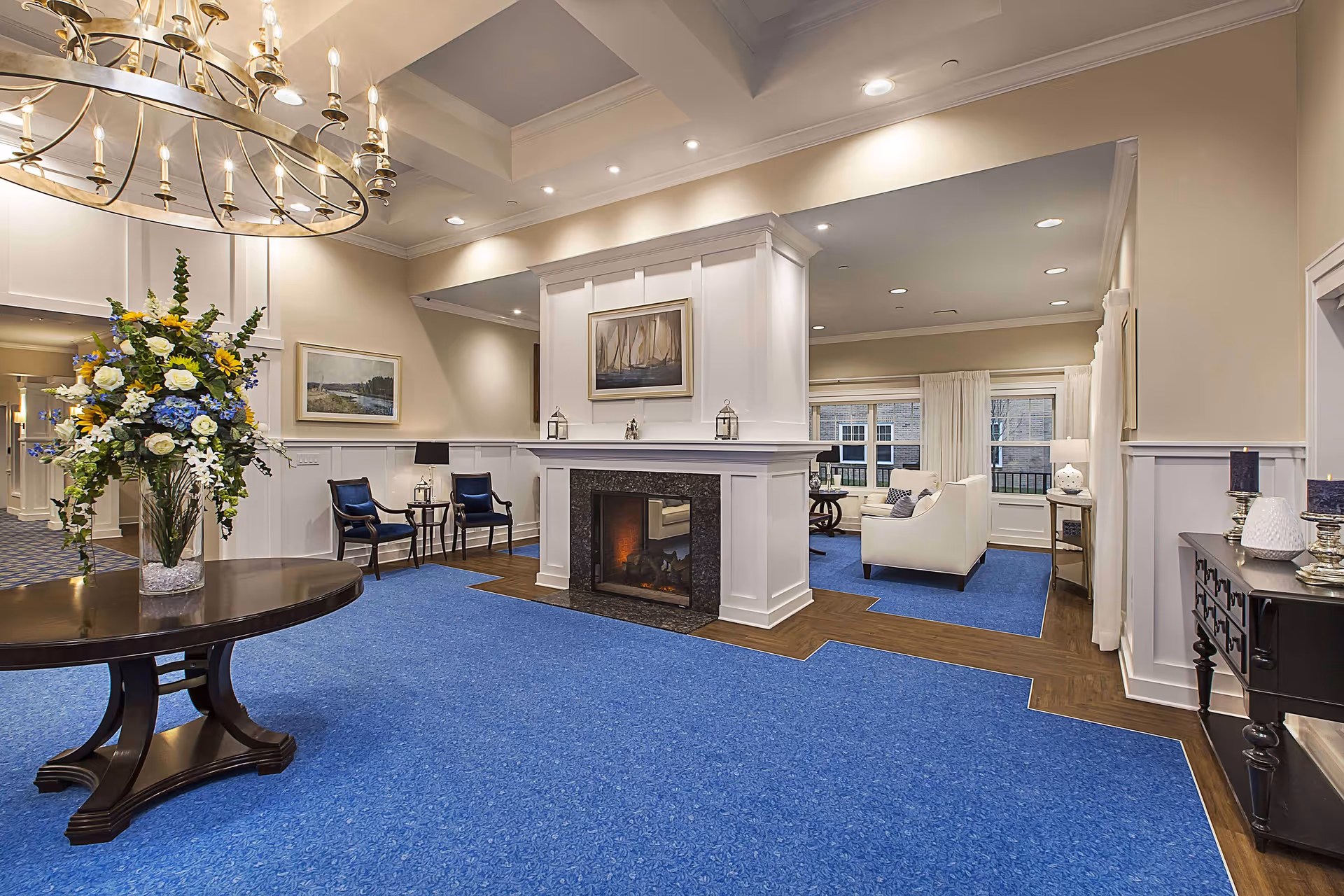 A spacious and elegantly decorated living room area in a senior living facility with blue carpet and wood flooring accents. The room features a large white fireplace with a painting above it, white sofas, and large windows with white curtains. There is a round wooden table with a tall floral arrangement and a chandelier hanging from the ceiling. Additional seating includes two blue upholstered chairs with a small table between them, and a black console table with decorative items on the right side.