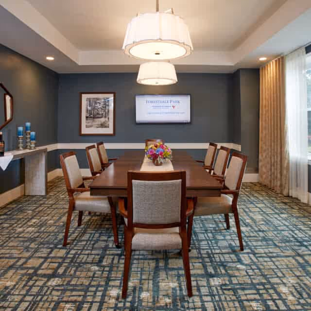 A formal dining room with a rectangular wooden table surrounded by eight upholstered chairs. Two large pendant lights hang above the table, and a floral centerpiece decorates the middle. The room has patterned carpet flooring, dark blue walls with white trim, a framed picture on one wall, and a flat screen displaying 'Forestdale Park Senior Living'. Large windows with beige curtains allow natural light into the room.