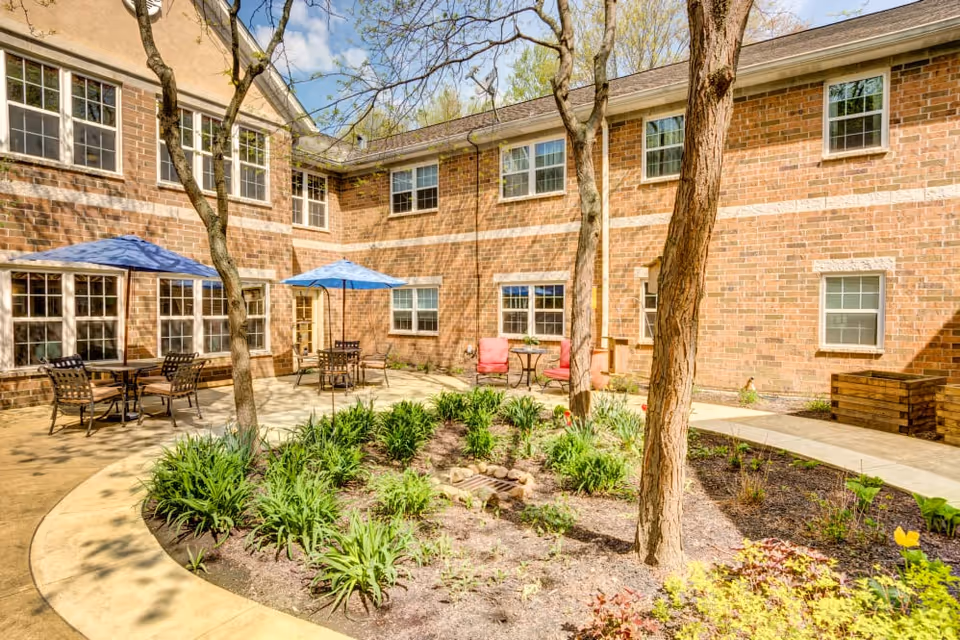 Outdoor courtyard area with brick building walls surrounding it, featuring several trees, garden beds with plants, and patio seating including tables with blue umbrellas and chairs, as well as a small table with two red cushioned chairs.