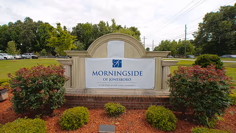 Outdoor view of a large stone and brick sign for Morningside of Jonesboro, surrounded by landscaped bushes and mulch with a parking lot and trees in the background under a cloudy sky.