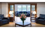 A cozy living room area with two black leather armchairs on either side, a wooden coffee table with a vase of pink flowers in the center, and a blue couch with patterned pillows in the background near a large window letting in natural light. Two table lamps on wooden side tables flank the couch.