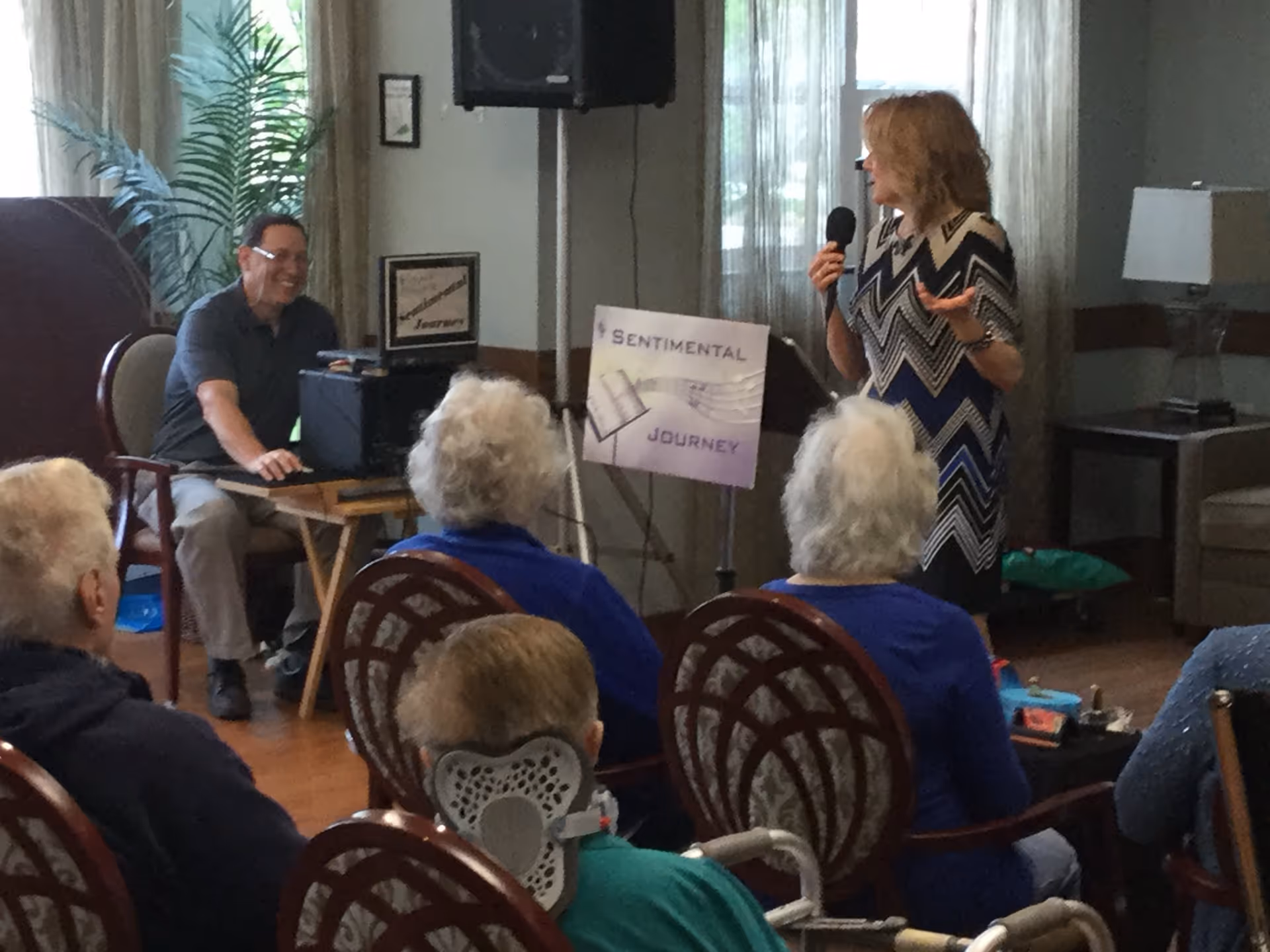 A woman holding a microphone is speaking to a seated audience of elderly people in a room. A man is sitting at a table with audio equipment and a speaker behind him. A sign next to the speaker reads 'Sentimental Journey'. The room has chairs, a lamp, and a plant near a window with curtains.