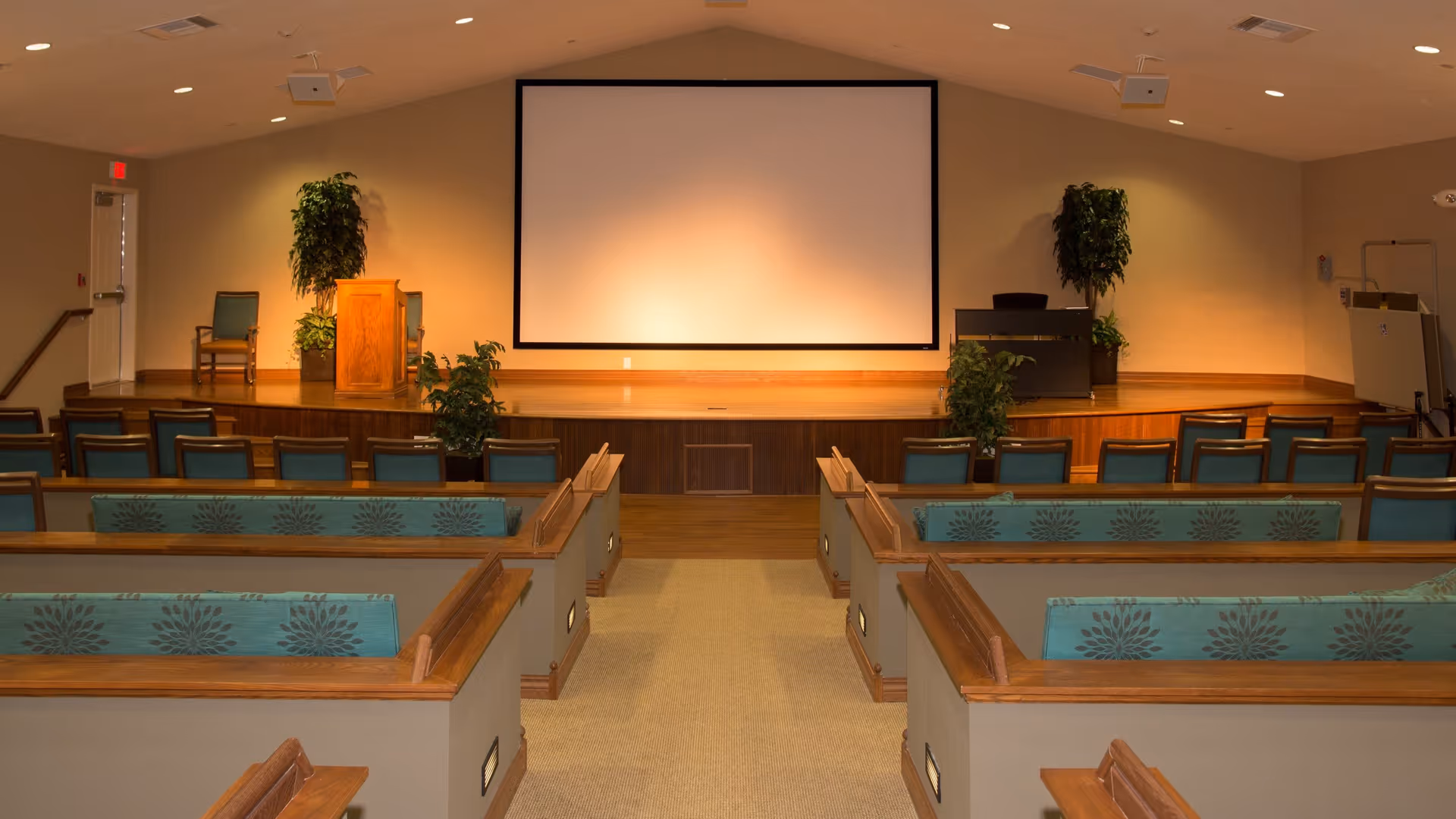 Interior view of a presentation or meeting room with rows of wooden pew-style seating with teal cushions facing a stage. The stage has a wooden podium, two potted plants, and a large blank projection screen on the wall behind it. The room is well-lit with recessed ceiling lights.