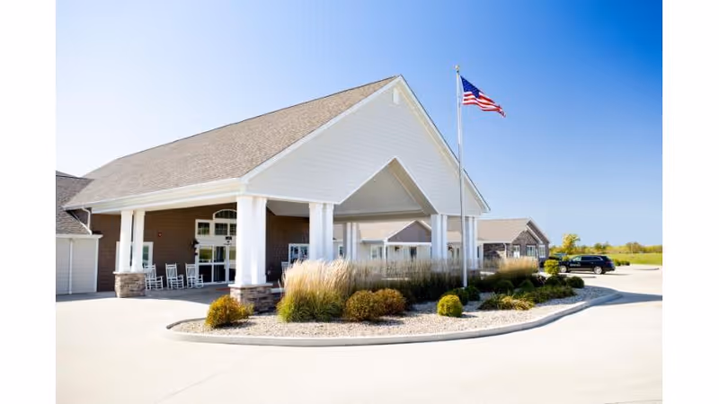 Entrance of a senior living facility with a covered porte-cochère, white columns, landscaping, and an American flag.