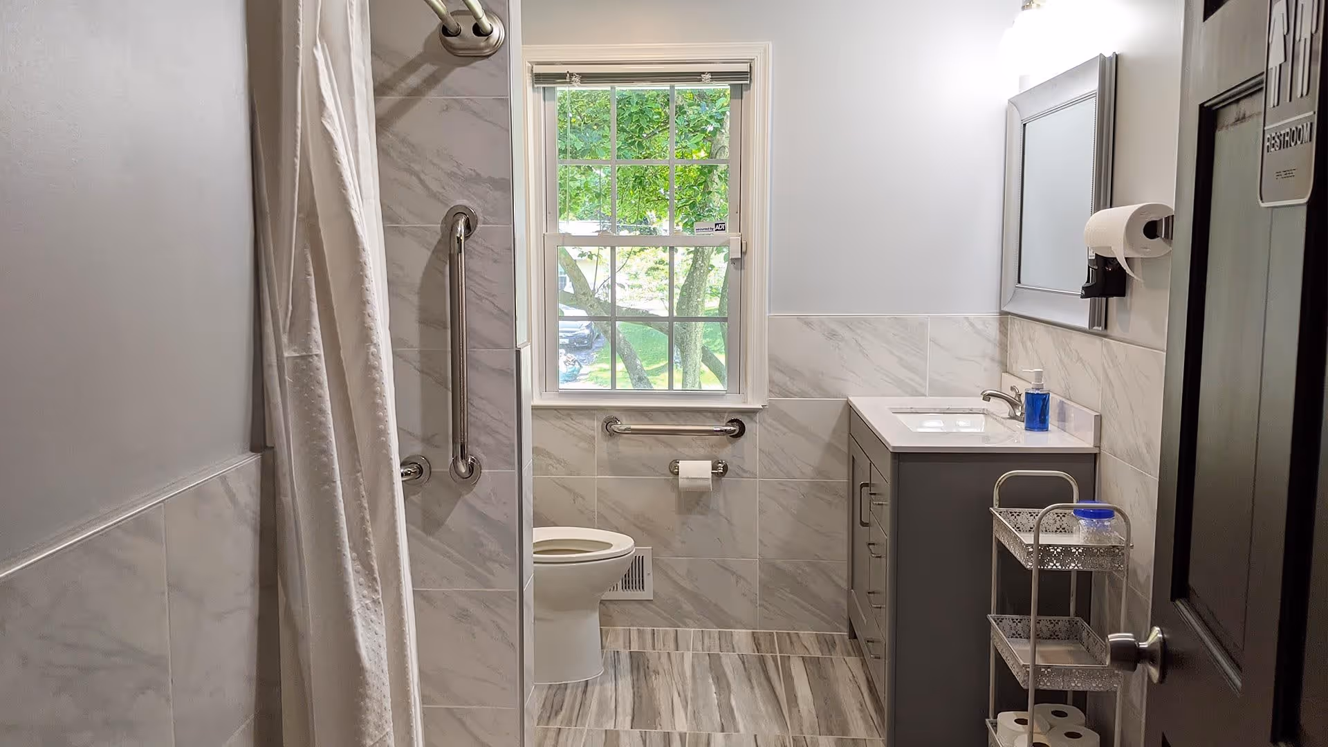 A clean and modern bathroom with a shower area on the left featuring a grab bar and white shower curtain. In the center, there is a toilet beneath a window with a view of green trees outside. On the right side, there is a gray vanity with a sink, a mirror above it, a soap dispenser, and a paper towel holder mounted on the wall. A small metal rack with toilet paper rolls and a container is next to the vanity. The door on the right has a sign that reads 'RESTROOM'.