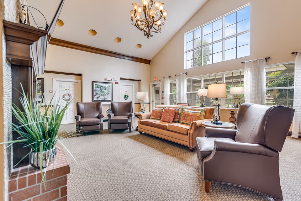 A bright and spacious living room with high vaulted ceilings and large windows letting in natural light. The room features a brown leather armchair, a brown sofa with colorful cushions, two additional brown armchairs, a side table with a lamp, a chandelier hanging from the ceiling, and a fireplace with a plant on the mantel. The walls are light-colored with wooden trim, and there are framed pictures and white curtains on the windows.