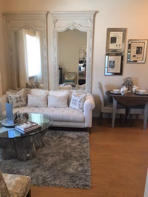 Cozy living area with a white tufted sofa in front of two tall ornate mirrors, a glass coffee table on a shag rug, and a small round dining table set against the wall.