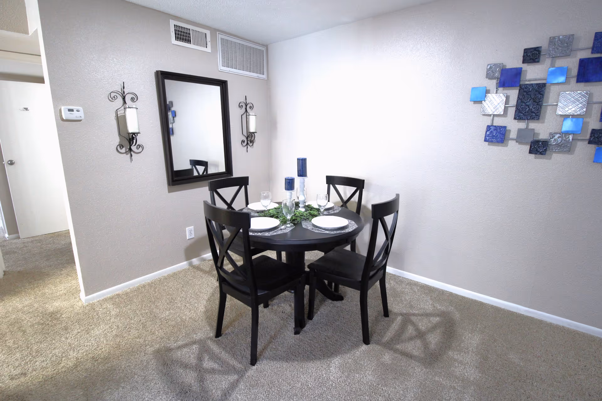 A small dining area with a round black table set for four with white plates, glasses, and blue candles in the center. The room has beige carpet and light-colored walls. A large mirror is mounted on one wall, flanked by two decorative candle holders. A modern blue and silver wall art piece is displayed on another wall.