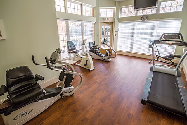 A small fitness room with exercise equipment including a treadmill, recumbent bikes, and an elliptical machine. The room has large windows with blinds, wood flooring, and a wall clock above the windows.