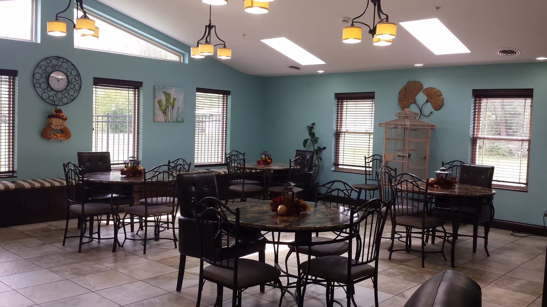 A bright dining room with multiple round tables and chairs arranged neatly. The walls are painted light blue with several windows featuring wooden blinds. There are decorative items on the walls including a clock, a painting, and metal leaf art. The ceiling has modern light fixtures and skylights providing natural light. The floor is tiled and there is a birdcage in the corner.