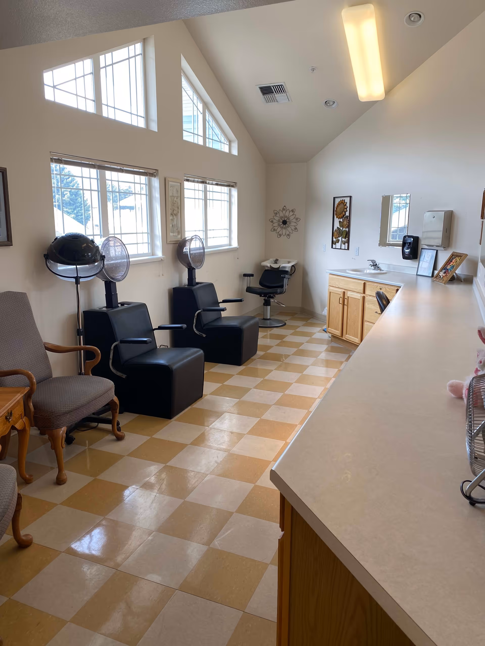 Interior view of a hair salon area in a retirement and assisted living facility with two black salon chairs under hair dryers, a washing station with a chair, a long countertop with a sink, and large windows letting in natural light.