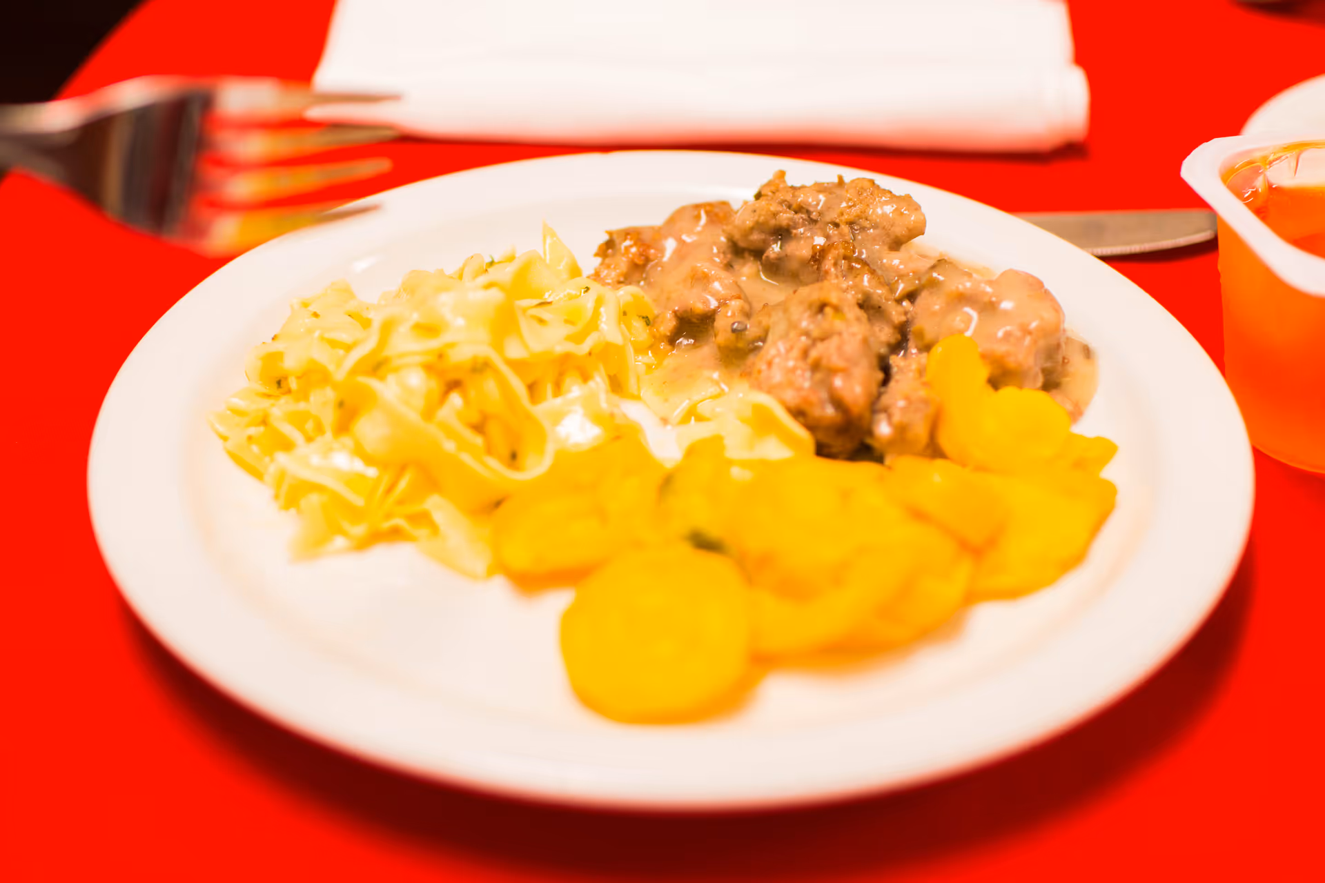 A white plate with a serving of egg noodles, ground meat with gravy, and cooked yellow squash on a red tablecloth. A fork and knife are placed near the plate, and a small cup of applesauce is visible on the right side.