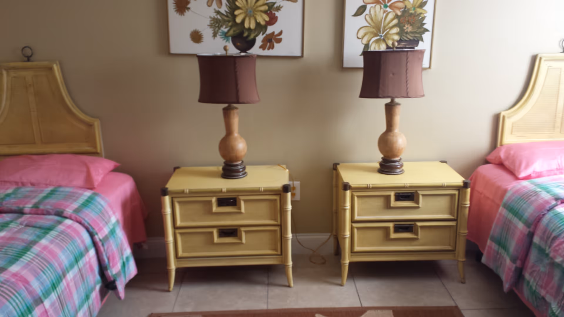 Two twin beds with pink bedding flank matching yellow nightstands topped with wooden lamps and floral artwork on the wall.