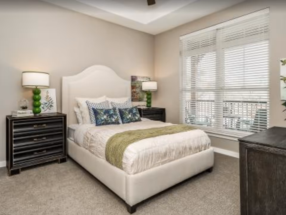 A neatly made bed with white bedding, green throw blanket, and decorative pillows in a bedroom. The room has beige walls, carpeted floor, two black nightstands with green lamps, a large window with white blinds, and a ceiling fan.