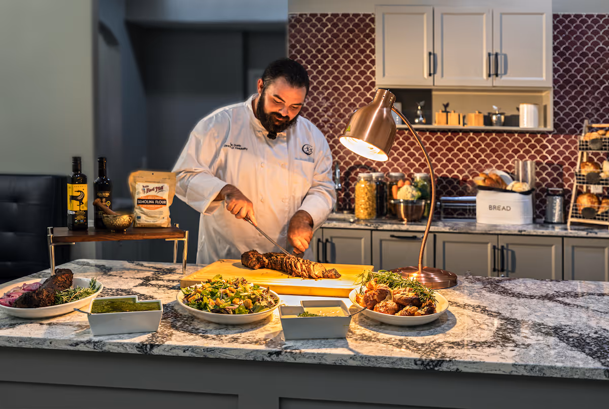 A chef in a white coat is slicing a piece of cooked meat on a wooden cutting board in a kitchen. The countertop in front of him has plates of food including salad, meat, and dipping sauces. The kitchen has gray cabinets, a red patterned backsplash, and various kitchen items like jars, bread, and bottles on the counter.