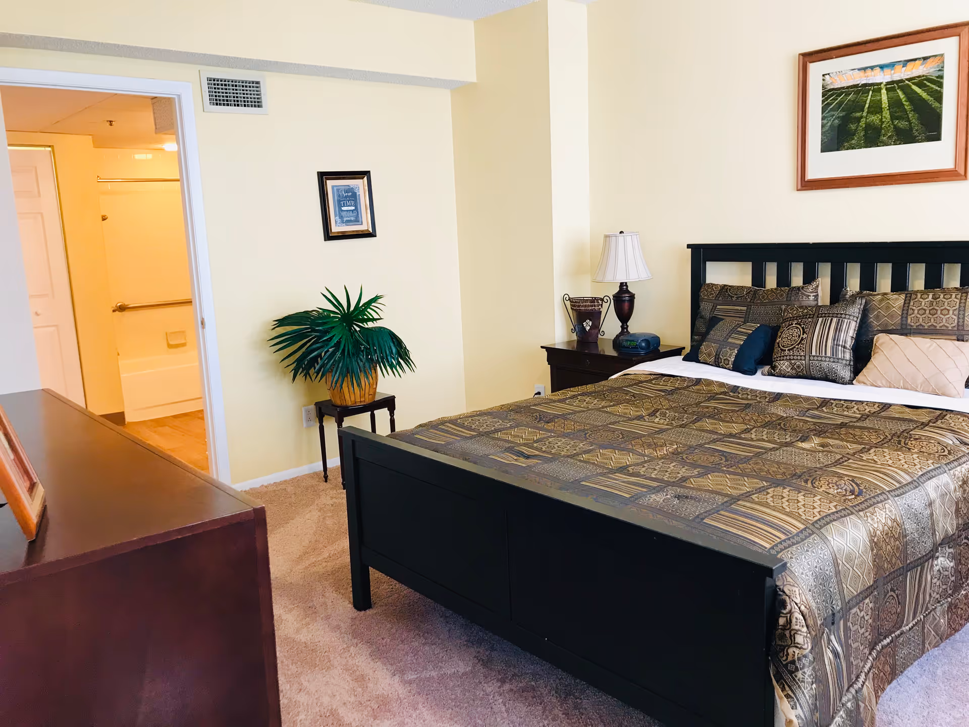 A bedroom with a black wooden bed frame and a patterned bedspread with multiple pillows. There is a nightstand with a lamp and a clock radio next to the bed. A framed picture hangs on the wall above the bed. To the left, there is a small table with a potted plant and a framed picture on the wall. An open door reveals a bathroom with a bathtub and grab bars.
