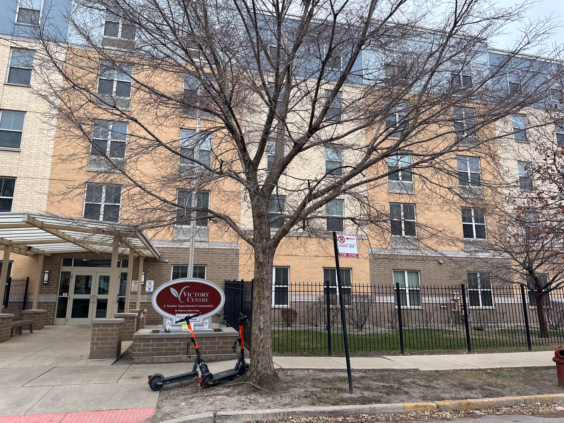 Front exterior of the Victory Centre supportive living building showing the entrance, facility sign, a leafless tree, and two parked scooters.