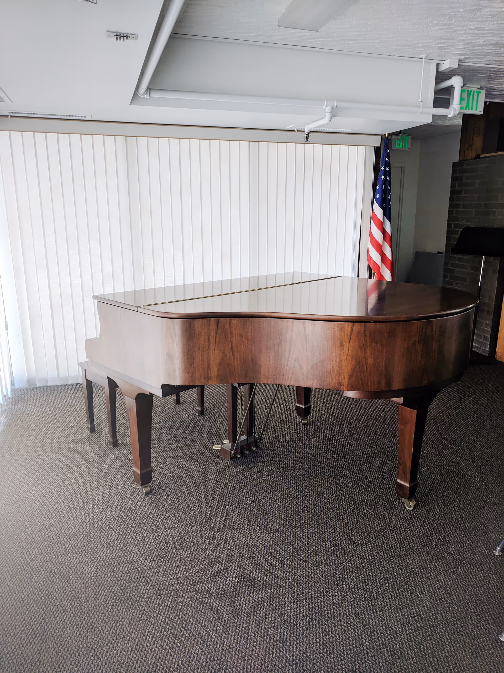 A wooden grand piano with a closed lid is placed on a carpeted floor in a room with vertical blinds covering the windows. An American flag stands in the corner near a doorway with an exit sign above it.
