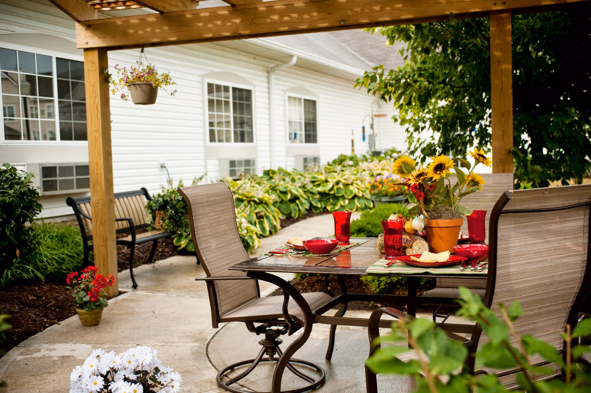 Outdoor patio area with a glass-top table set for a meal, featuring red glasses, plates, and a centerpiece with sunflowers. The patio is covered by a wooden pergola and surrounded by green plants and flowers. A white building with windows is visible in the background.