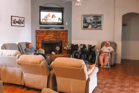 A cozy living room area in a senior living facility with several beige armchairs arranged around a brick fireplace. A television is mounted above the fireplace, and two elderly individuals are seated in the room, one in an armchair and the other in a wheelchair. There are multiple wheelchairs lined up against the wall, a painting hanging on the wall, and a sign that reads 'thankful'.