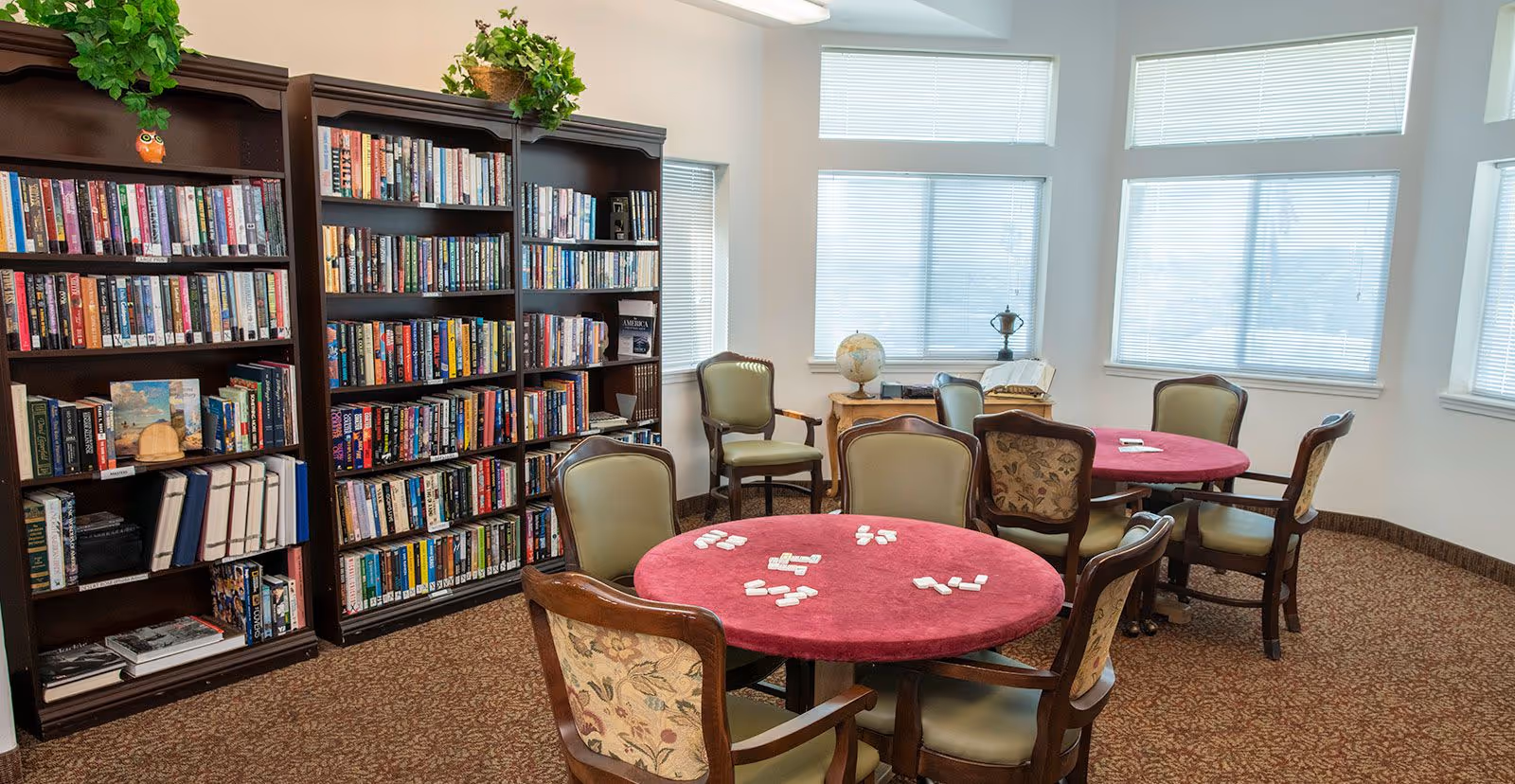 A cozy room with two round tables covered with red tablecloths, surrounded by cushioned chairs. Several white game pieces are arranged on the tables. Against the wall are tall dark wooden bookshelves filled with books and decorated with green plants. Large windows with blinds allow natural light to fill the room.