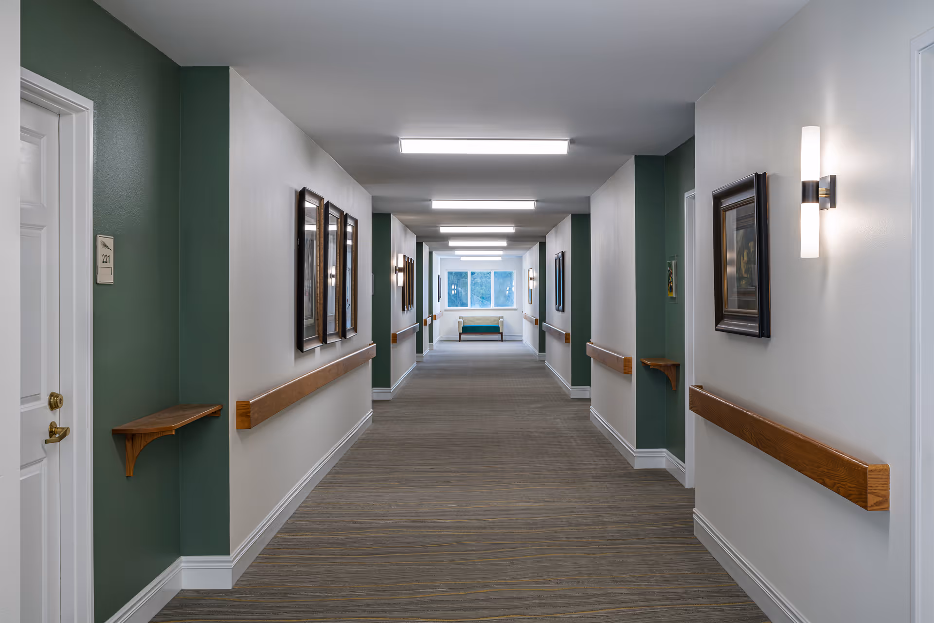 A long, well-lit hallway in a senior living facility with green and white walls, wooden handrails on both sides, framed artwork, and a bench at the far end near a window.