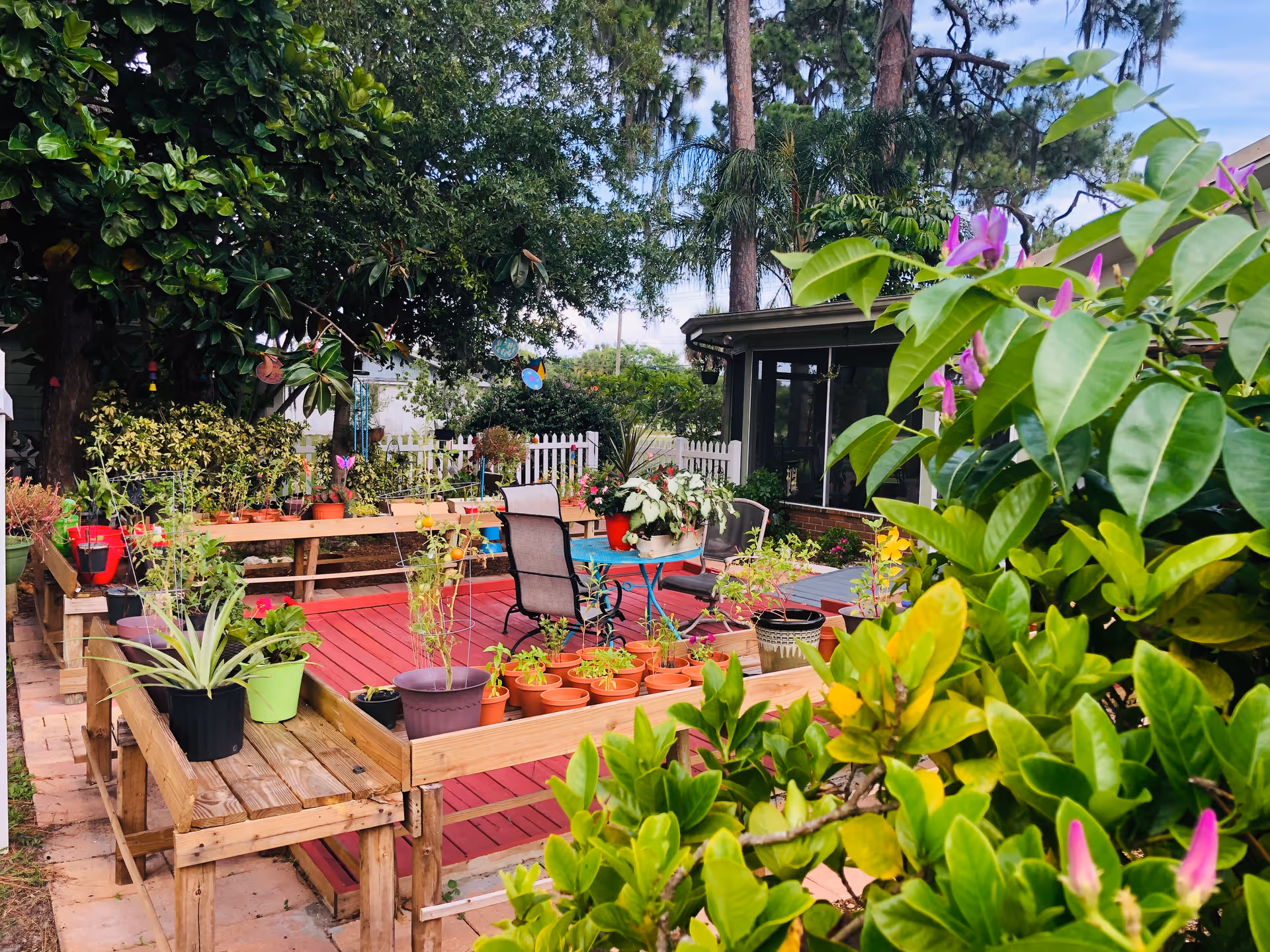 A colorful backyard deck filled with potted plants, patio chairs and a small table surrounded by lush greenery and a white picket fence.