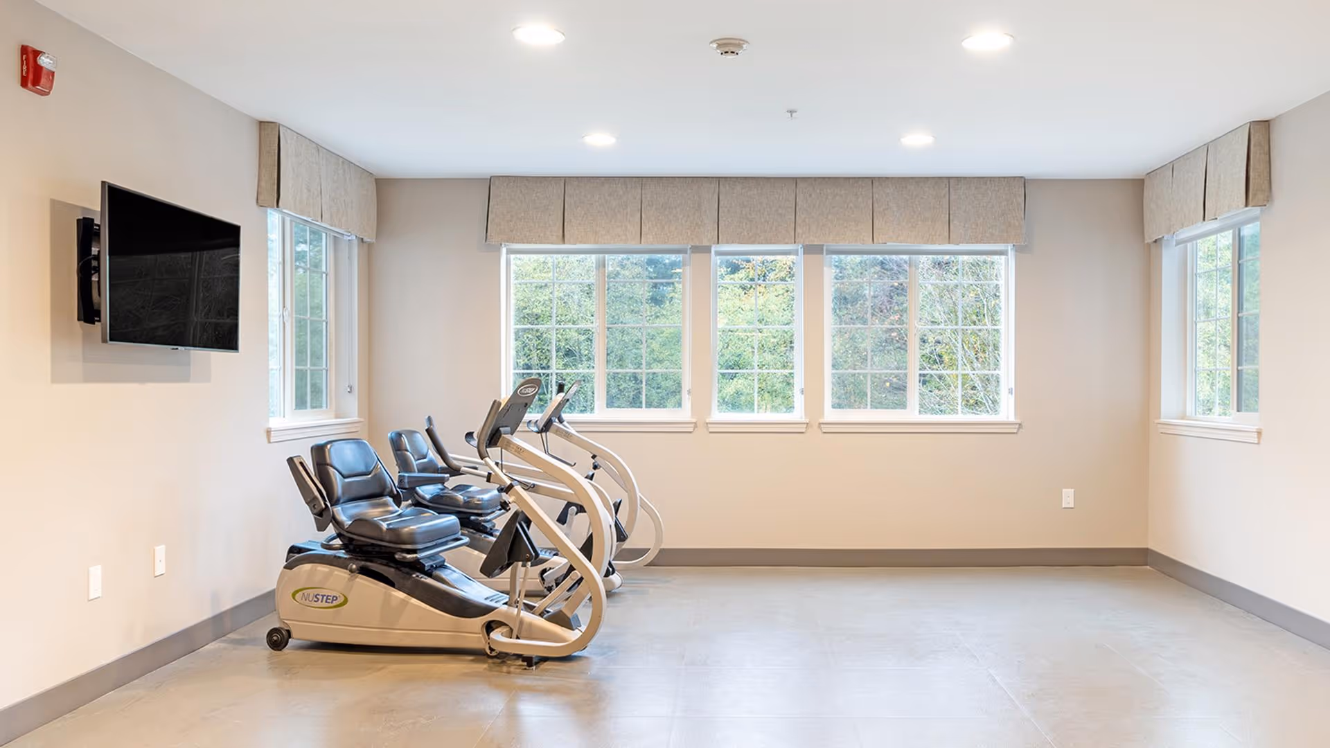 A bright exercise room with three NuStep recumbent cross trainers positioned near three large windows with beige valances. A flat-screen TV is mounted on the left wall. The room has light-colored walls and flooring.