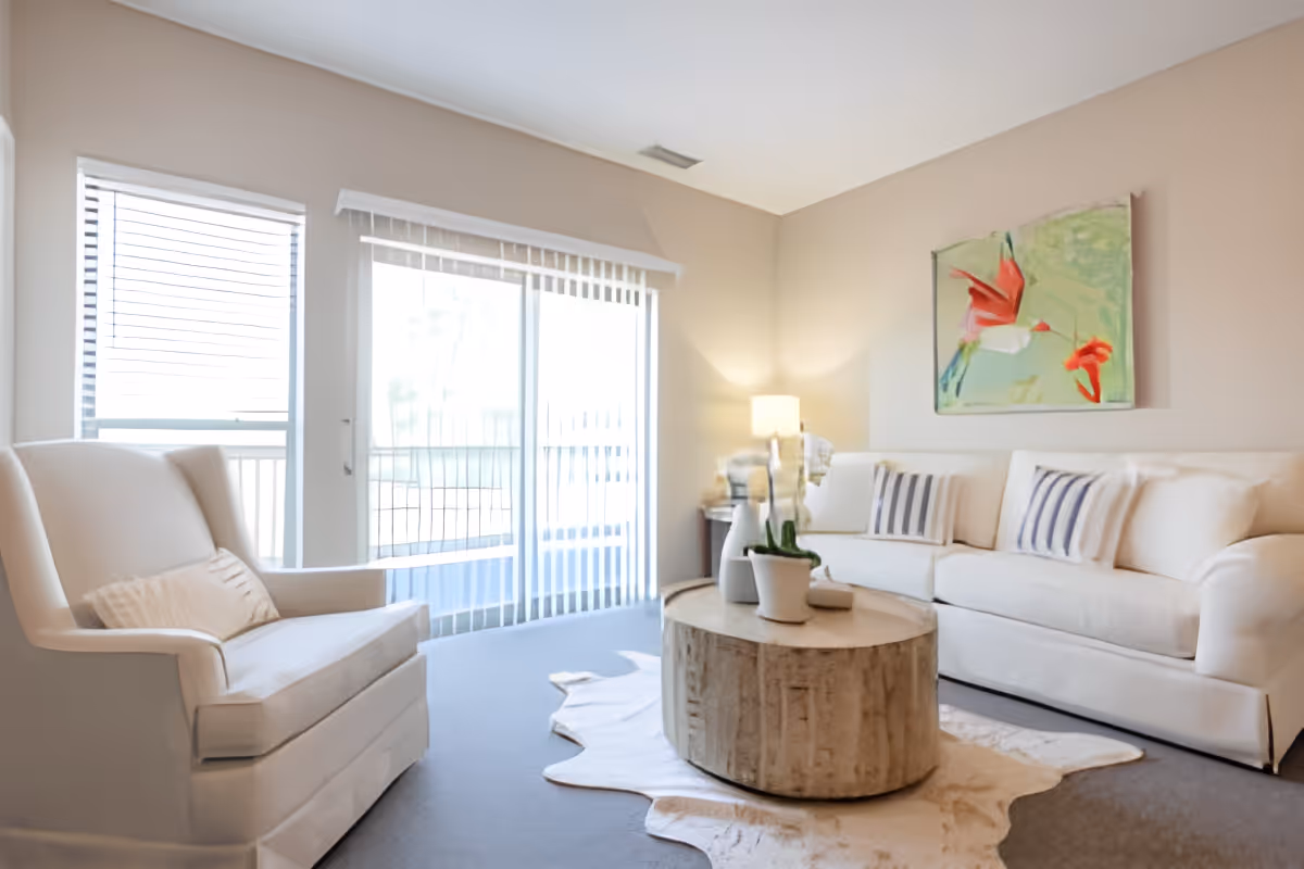 Bright living room with a white sofa and armchair, round wooden coffee table, and sliding glass doors to a balcony.
