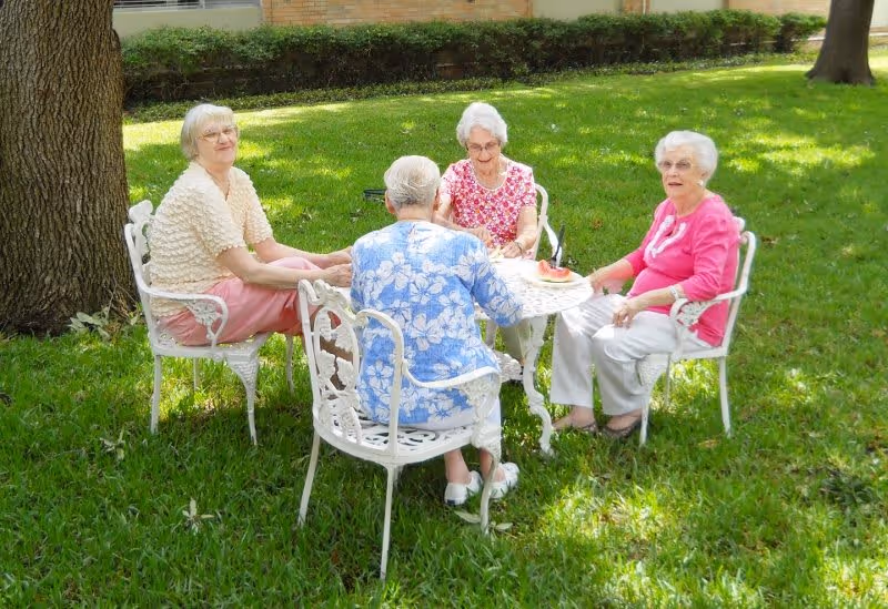 Four elderly women sitting around a white metal patio table on a grassy lawn under trees.