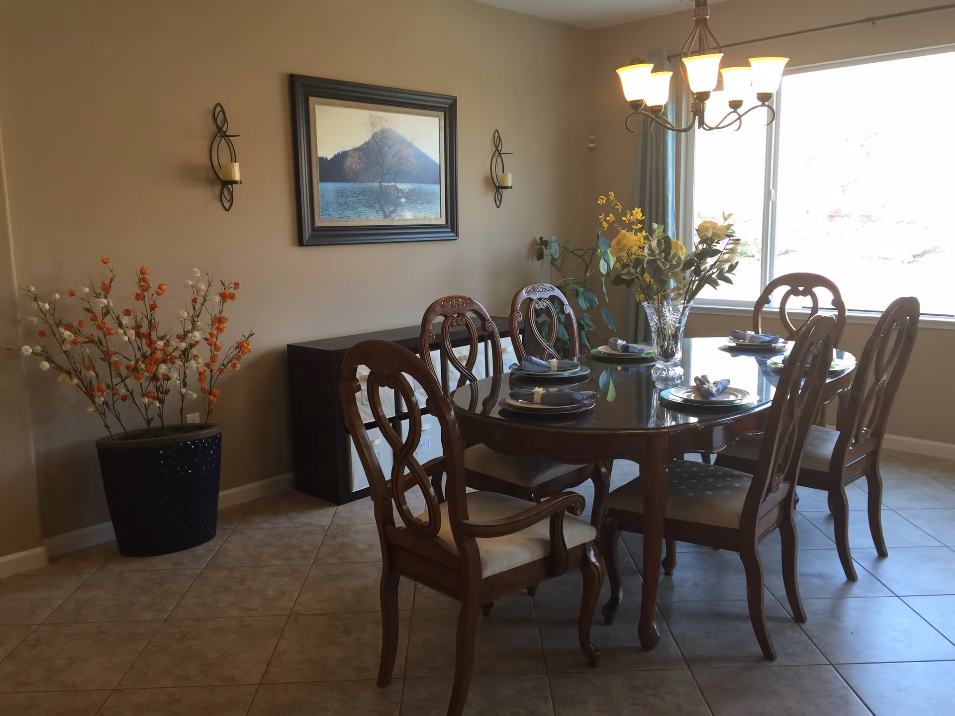 A dining room with a wooden dining table set for six people. The table has a glass top and is decorated with a vase of yellow flowers in the center. There are six wooden chairs with cushioned seats around the table. The room has beige walls, a large window letting in natural light, a chandelier hanging above the table, a framed landscape painting on the wall, and a large potted plant with orange and white flowers in the corner.