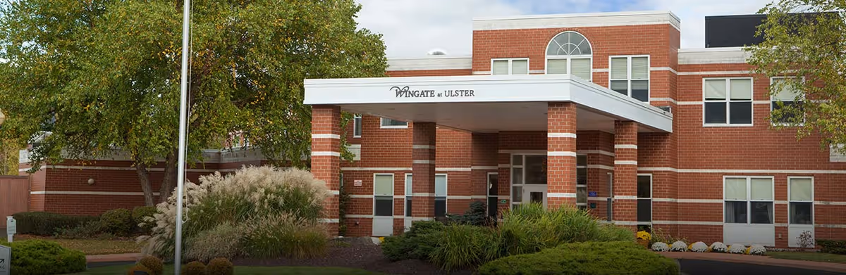 Front entrance of a red brick senior living building with a white canopy reading 'Wingate at Ulster' and landscaped greenery.