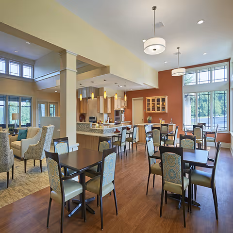 A bright and spacious dining area in Legacy Place Cottages featuring multiple tables with patterned chairs, a kitchen area with pendant lighting and granite countertops, large windows letting in natural light, and a cozy seating area with armchairs in the background.