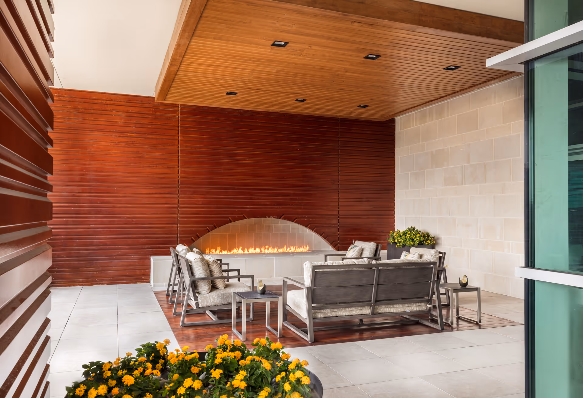 A modern indoor lounge area with cushioned chairs and a sofa arranged around a long, narrow fireplace set into a red wooden wall. The ceiling above the seating area is wooden with recessed lighting. There are yellow flowers in planters in the foreground and near the seating area. The floor is tiled in light colors, and there is a large glass window on the right side.