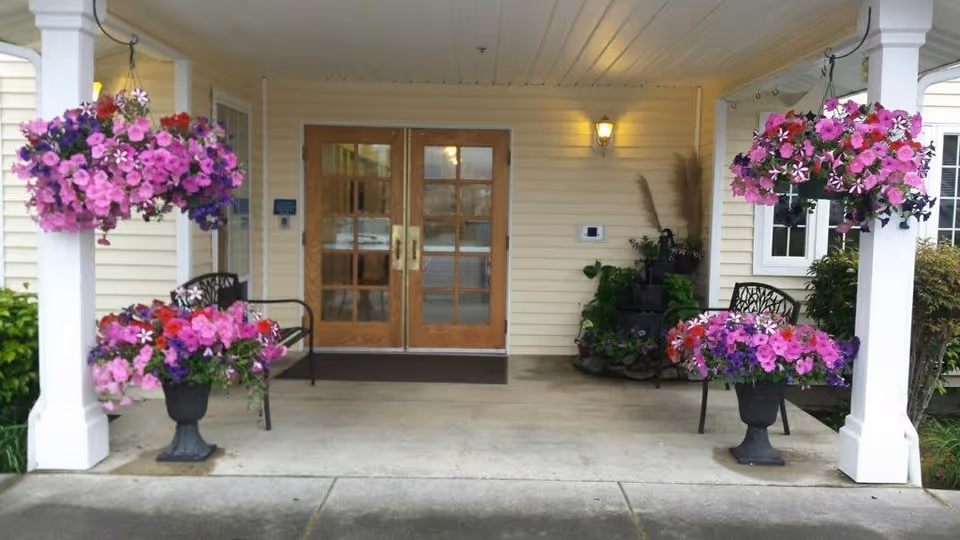 Entrance area of a senior living facility with double wooden doors featuring glass panels. The porch is decorated with hanging baskets and planters filled with vibrant pink, purple, and red flowers. Two black metal benches are placed on either side of the entrance under a covered porch with white pillars and a wall-mounted light fixture.