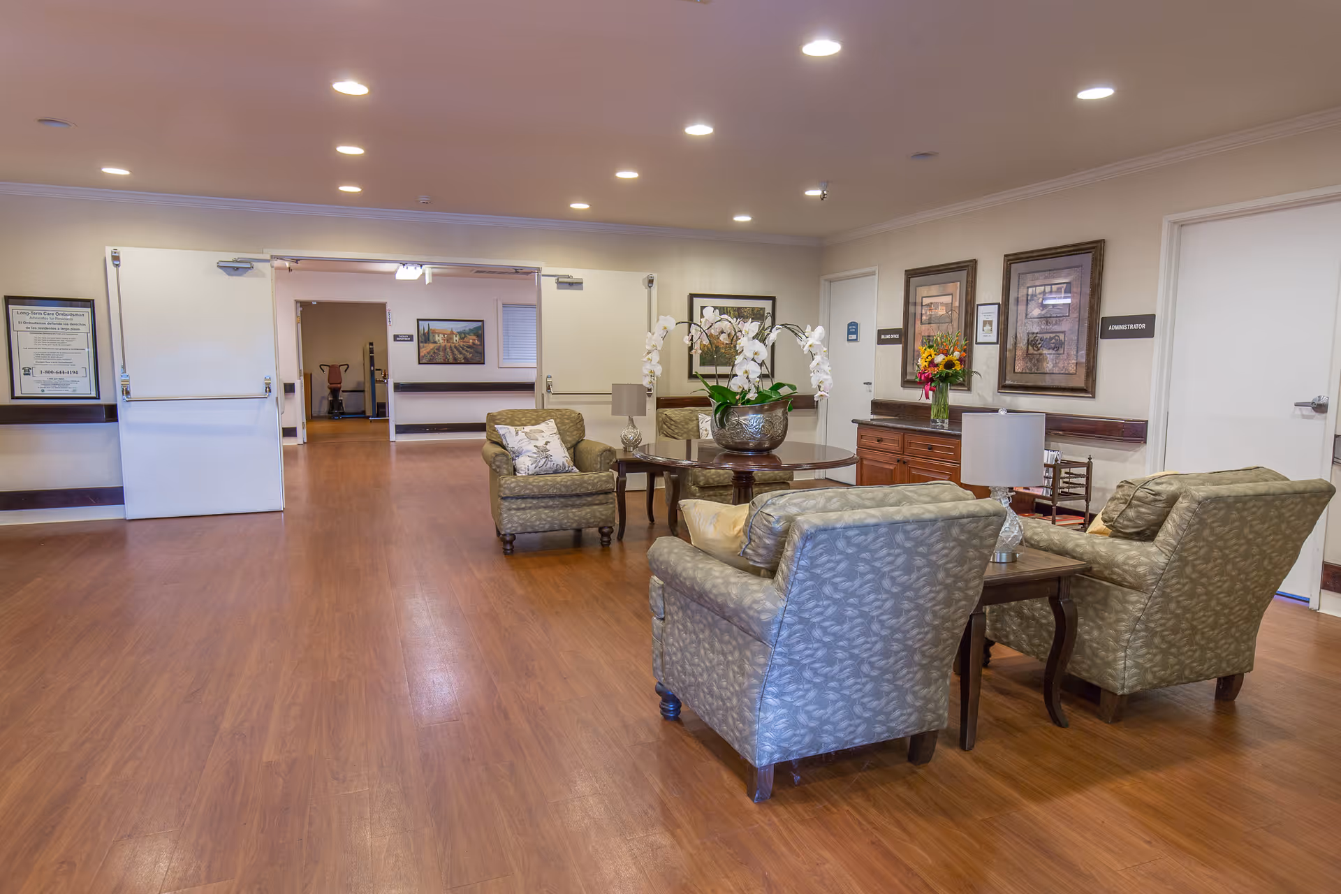 A spacious and well-lit common area in a senior living facility with wooden flooring, several upholstered armchairs arranged around a round wooden table with a large floral arrangement, framed artwork on the walls, and doors labeled 'Administration' and other rooms.
