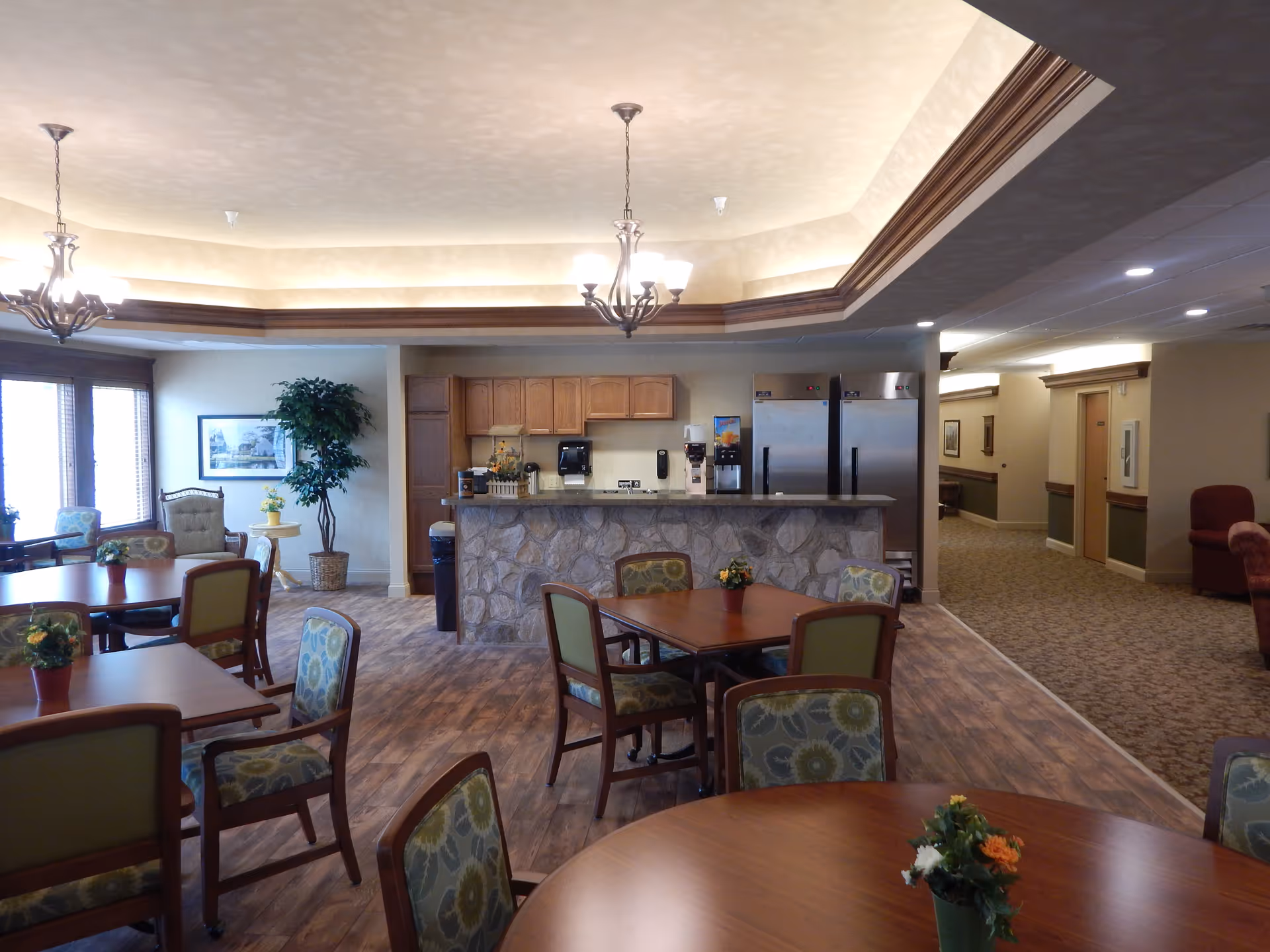 A spacious dining area in a senior living facility featuring multiple wooden tables with floral-patterned chairs. The room has a stone-faced counter with a coffee machine and two large stainless steel refrigerators behind it. The ceiling has decorative lighting fixtures and crown molding. There are large windows on the left side letting in natural light, and a hallway extends to the right.