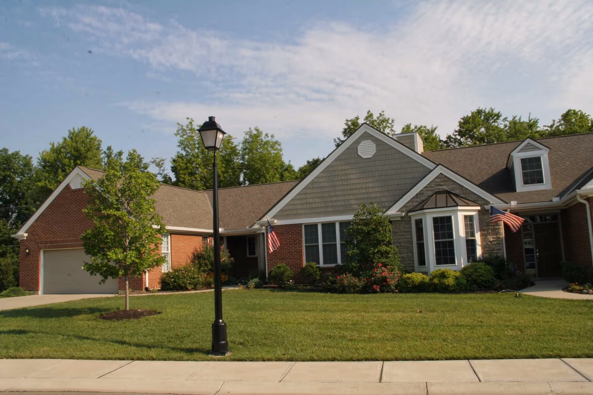Front exterior of a single-story brick and siding senior living building with a manicured lawn, lamp post, and American flags.