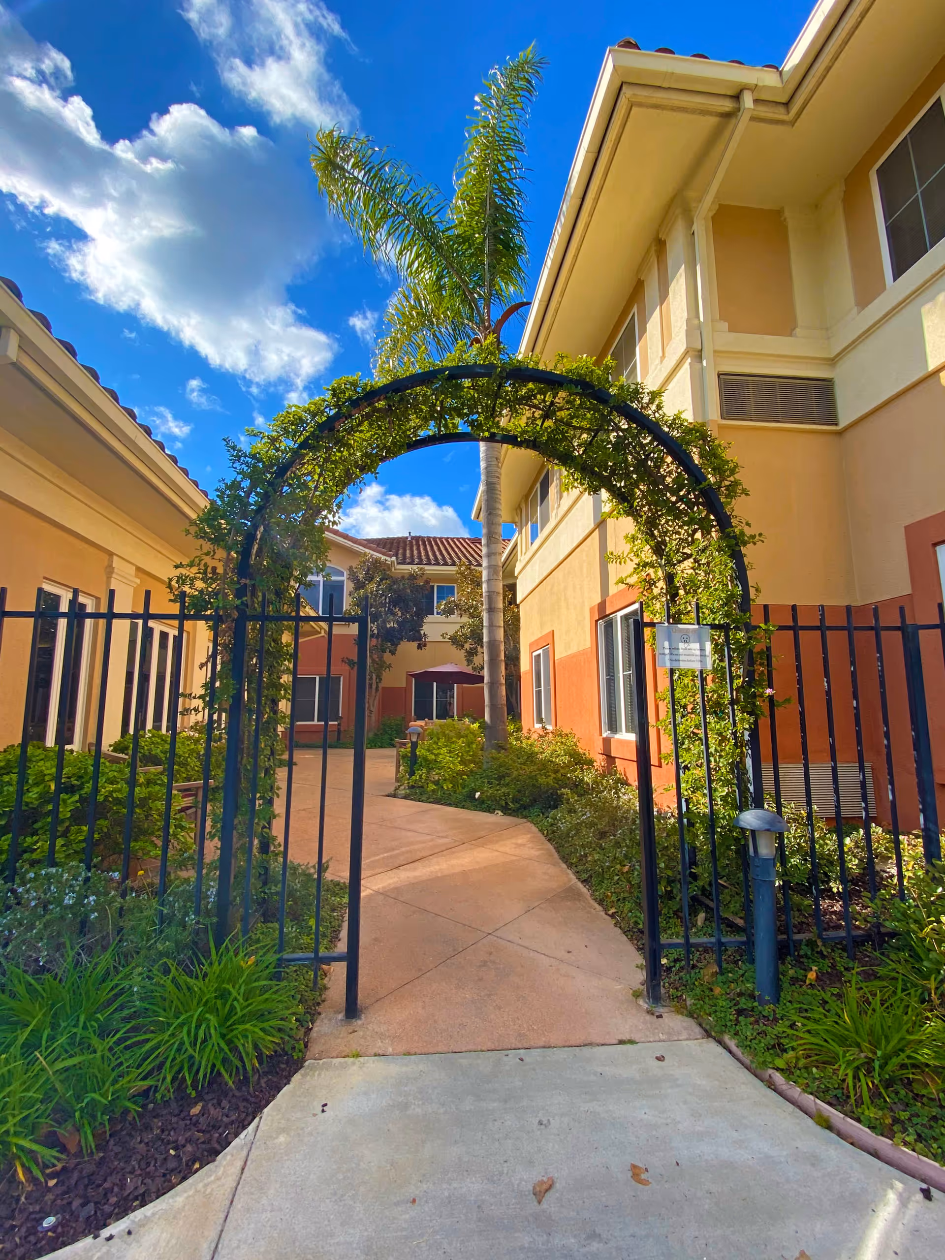 Wrought-iron arched gate leading into a landscaped courtyard between two beige-and-peach senior living buildings under a blue sky.