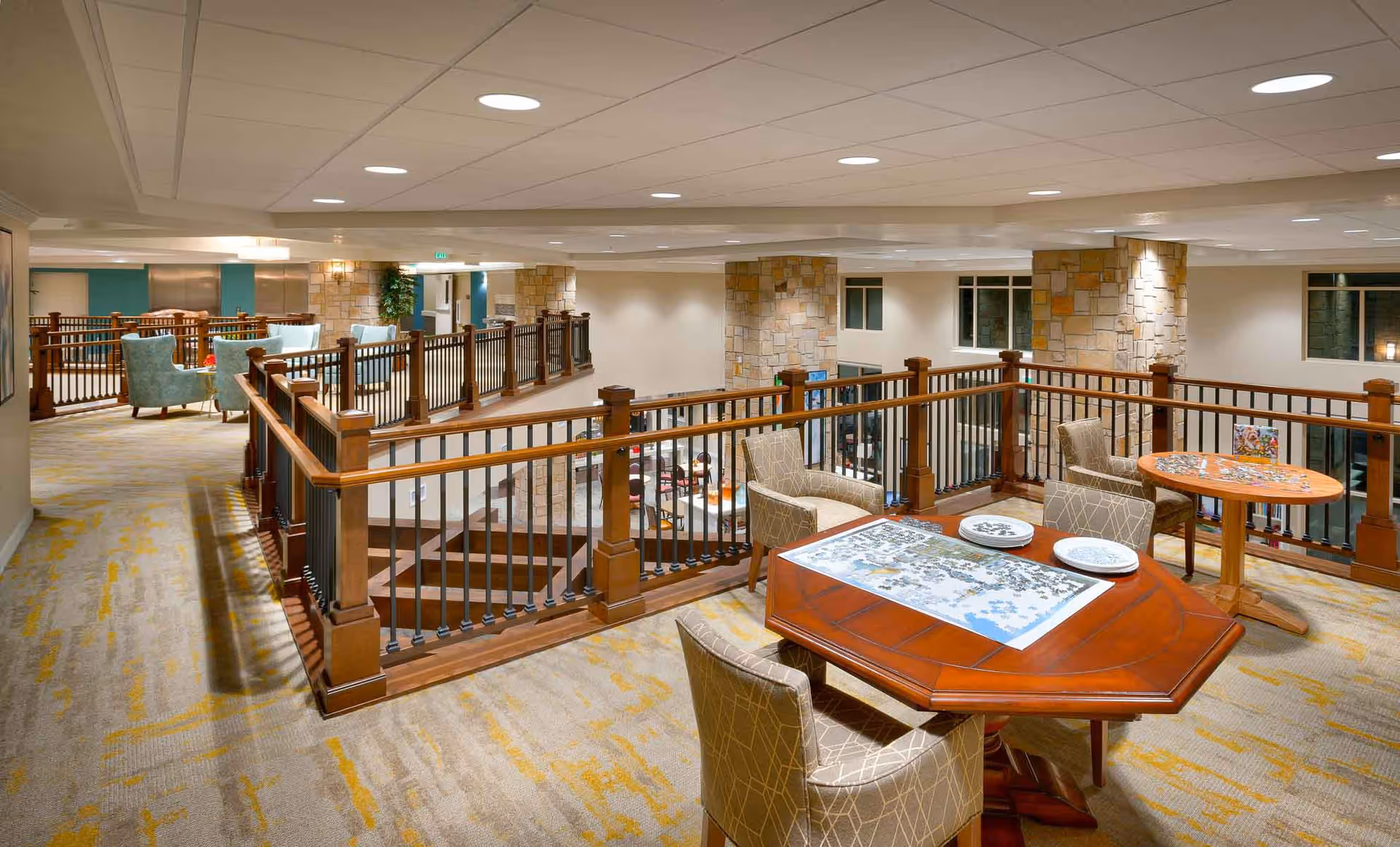 Interior view of a senior living facility common area with a puzzle table and chairs in the foreground, overlooking a lower level seating area with stone pillars and large windows. The space is well-lit with recessed ceiling lights and features carpeted floors and wooden railings.
