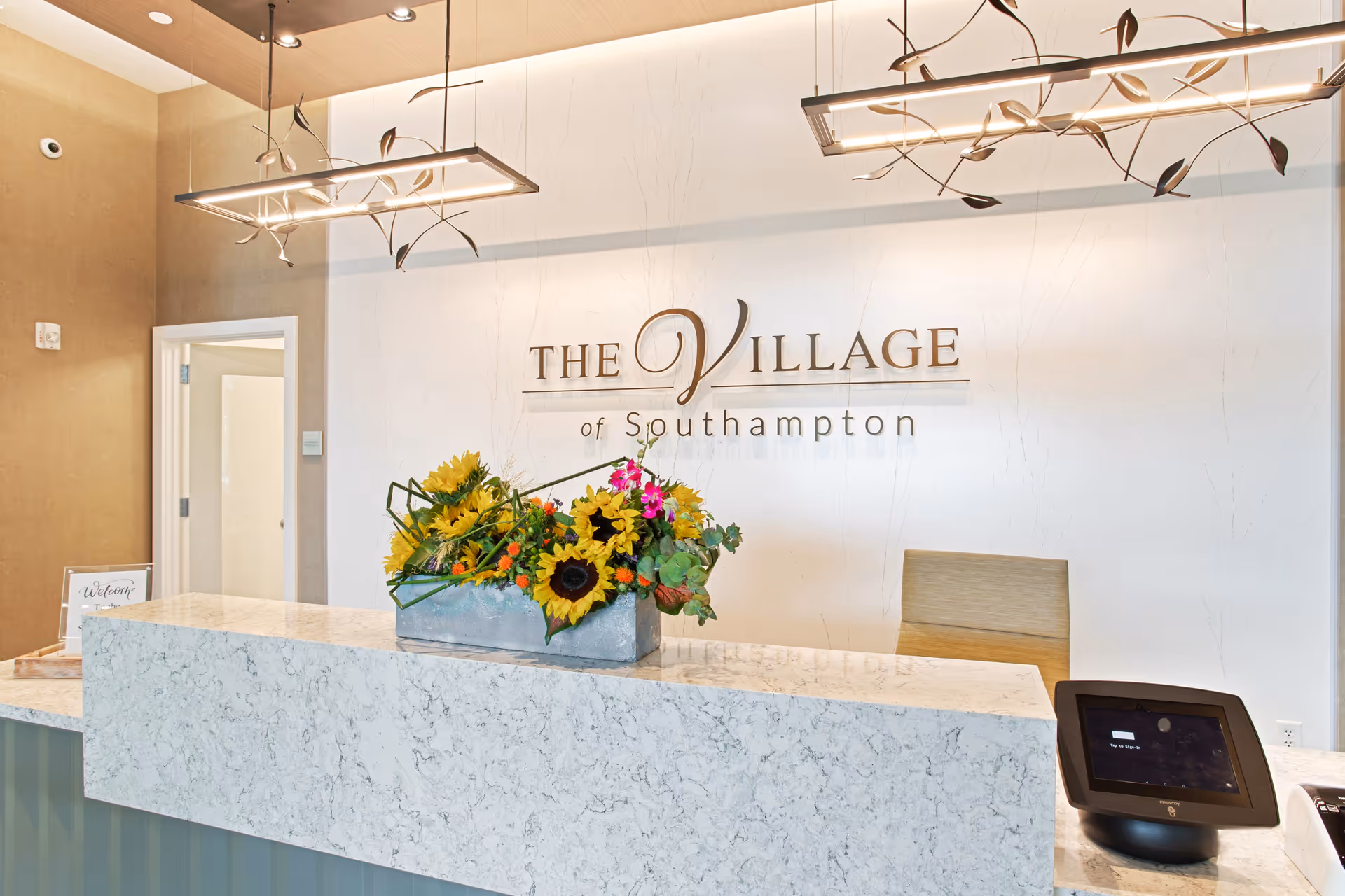 Reception desk area at The Village of Southampton with a marble countertop, a floral arrangement featuring sunflowers and other flowers, modern hanging light fixtures, a chair behind the desk, and a digital device on the counter. The facility name is displayed on the wall behind the desk.