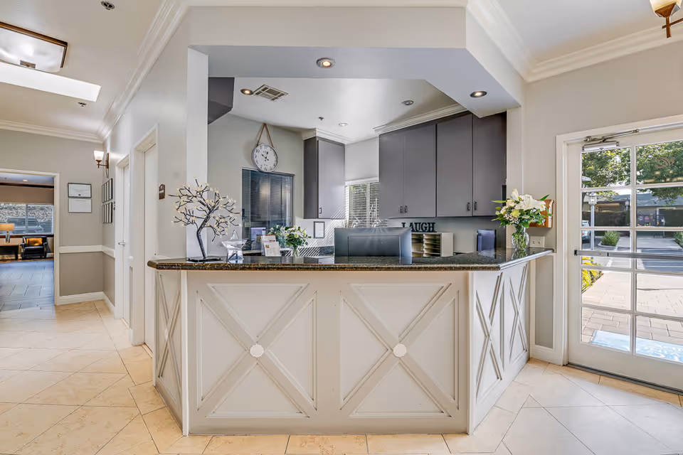 Reception desk area inside a senior living facility with a granite countertop, decorative items including a small tree sculpture and flower vases, gray cabinets behind the desk, a wall clock, and a glass door leading outside.