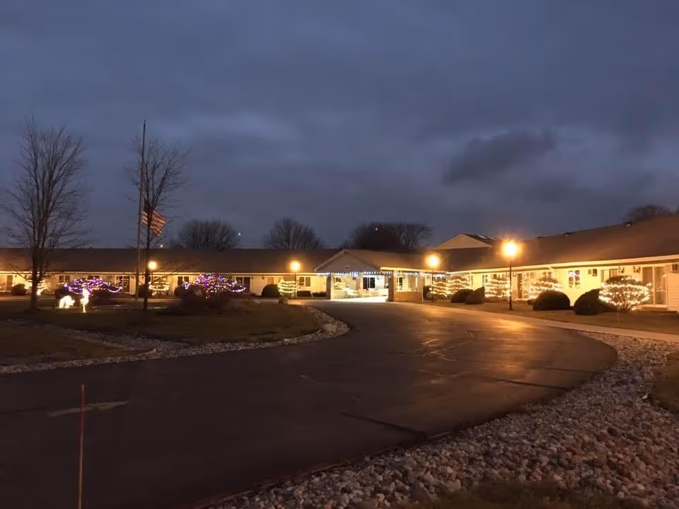 Front exterior of a single-story senior living building at dusk, decorated with holiday lights around bushes and along a circular driveway.