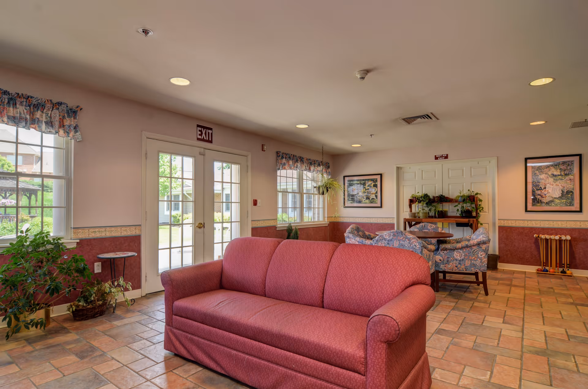 Interior view of a senior living facility common area with a red sofa in the foreground, a table with floral upholstered chairs, potted plants, framed artwork on the walls, and large windows with floral valances letting in natural light.