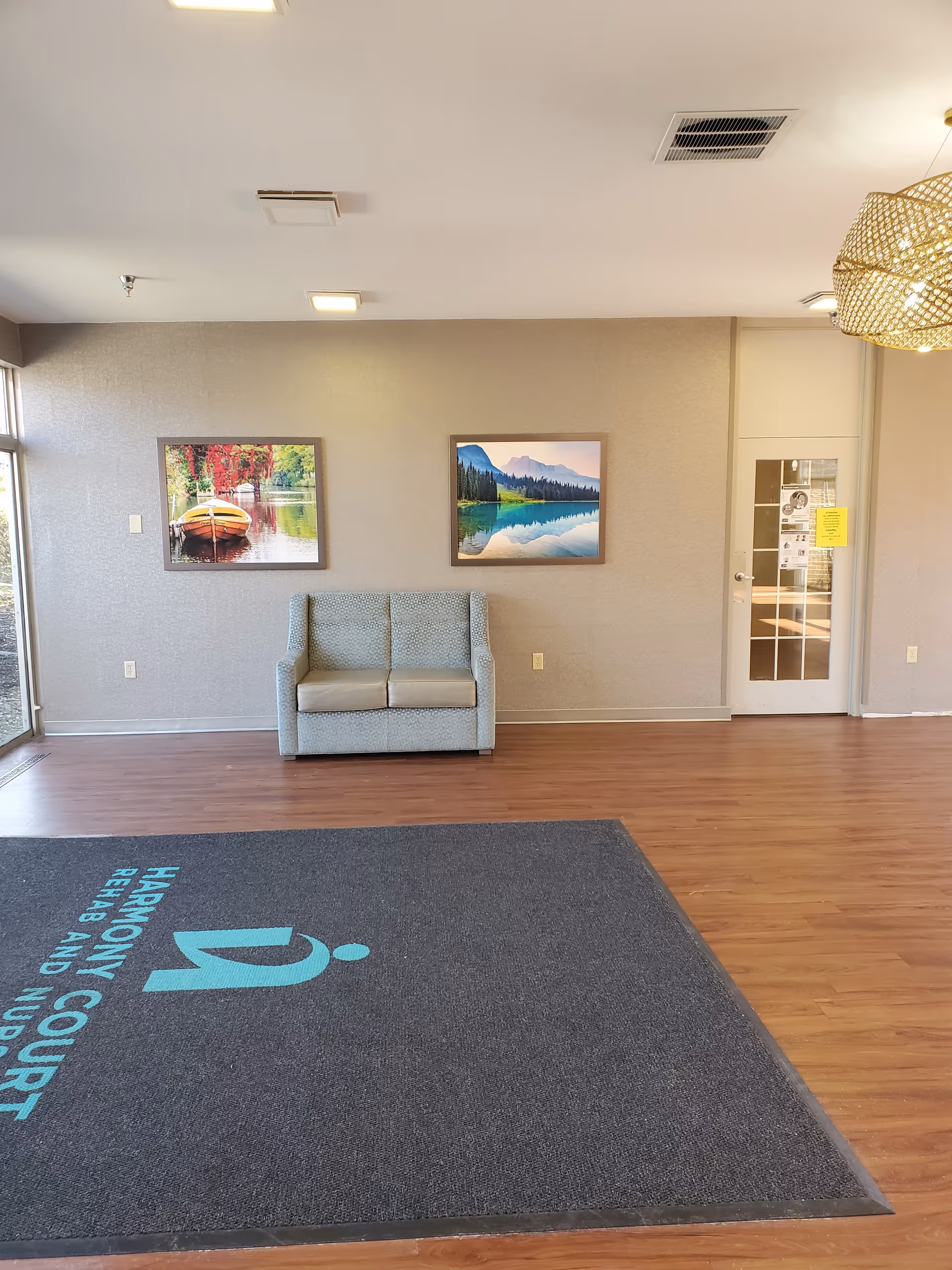 Interior view of a facility lobby with a small two-seater sofa against a beige wall. Two framed landscape paintings hang above the sofa. A large floor mat with the text 'Harmony Court Rehab and Nursing' is placed on the wooden floor. A glass door with notices is visible on the right side, and a decorative light fixture hangs from the ceiling.