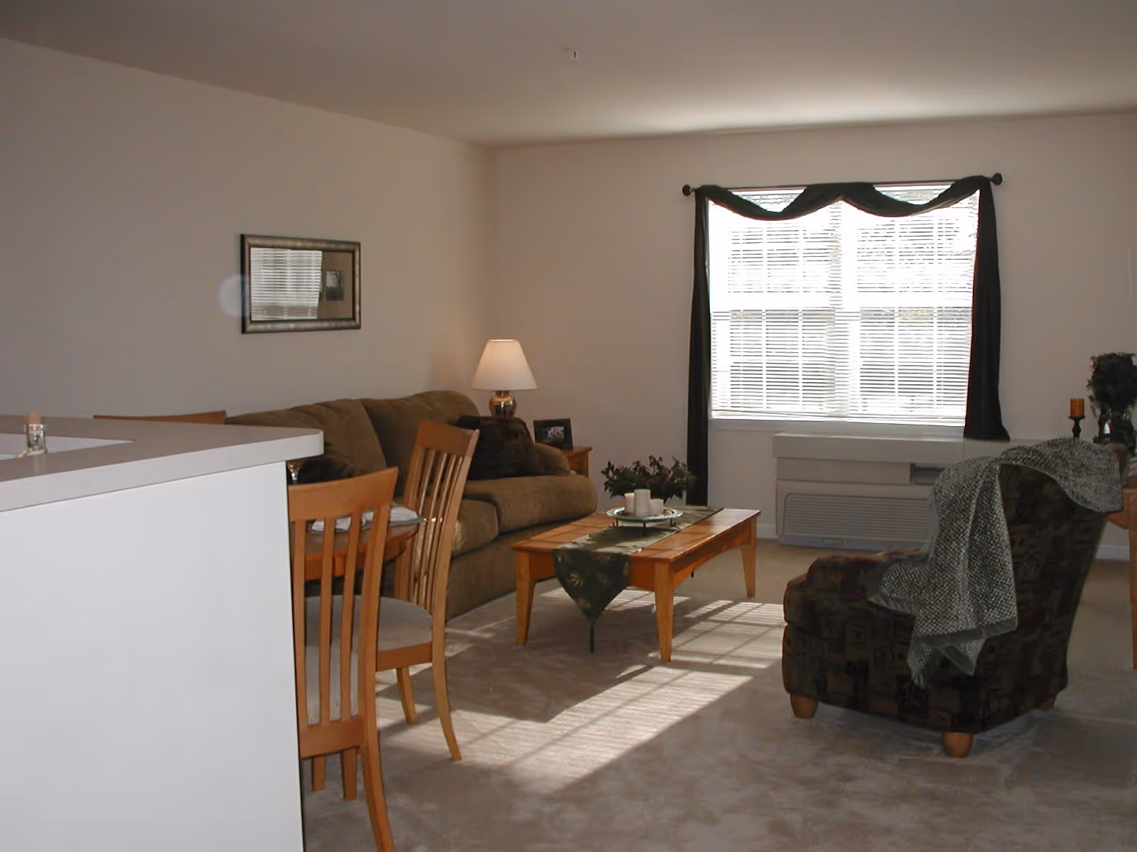A cozy living room with a brown sofa, a wooden coffee table with candles and a green table runner, a patterned armchair with a gray throw draped over it, a wooden dining table with chairs, a lamp on a side table, and a large window with blinds and dark curtains letting in natural light.