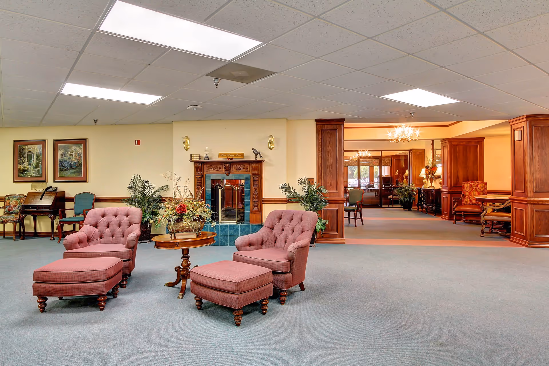 A spacious living room area in a senior living facility with two upholstered armchairs and matching ottomans in front of a wooden fireplace with blue tiles. A round wooden table with a floral arrangement is placed between the chairs. The room has carpeted floors, beige walls, framed artwork, and several potted plants. In the background, there is a hallway with additional seating and wooden paneling.