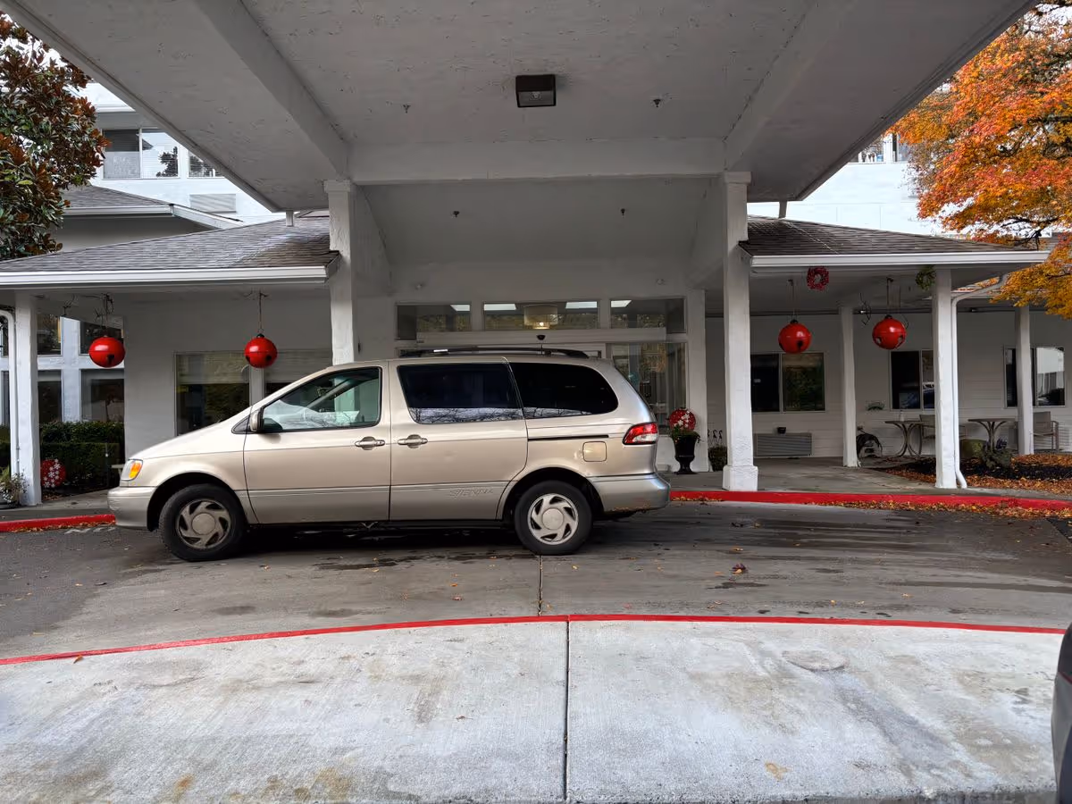 A beige minivan parked under the covered entrance/porte-cochere of a white senior living building with columns and red hanging decorations.