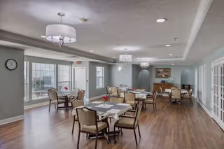 A spacious dining room with several tables and chairs arranged neatly. Each table has a small vase with red flowers. The room features large windows allowing natural light, light blue walls, wooden flooring, and modern ceiling lights. There is a clock on one wall and a sideboard with a framed picture above it in the background.